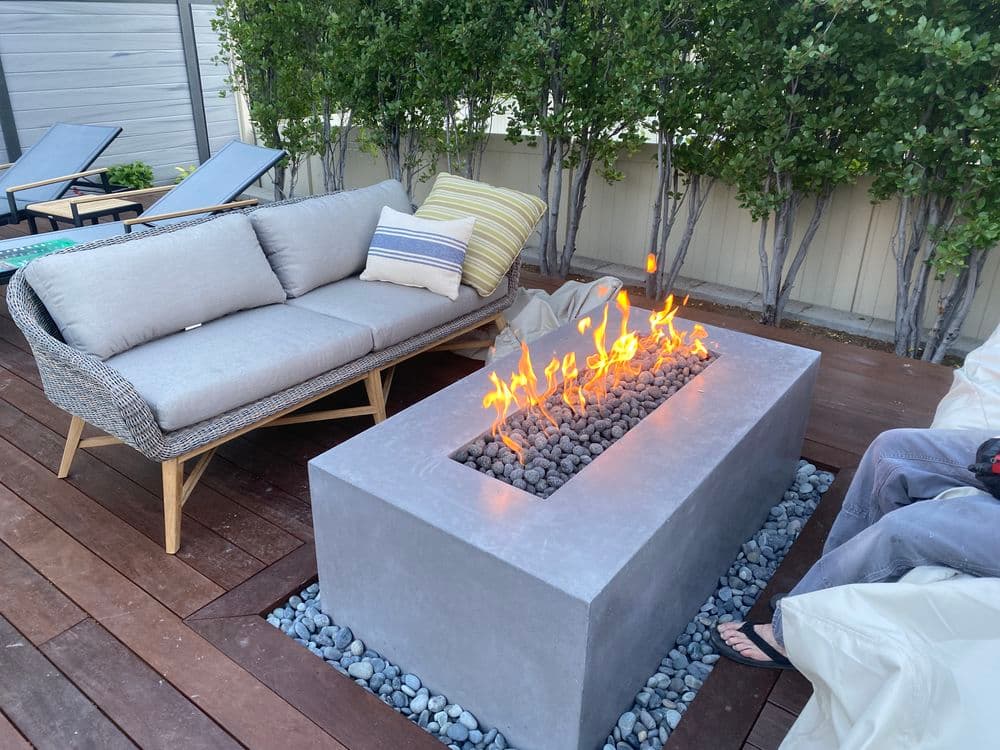 Modern outdoor seating area with a concrete fire pit and flames, surrounded by greenery.