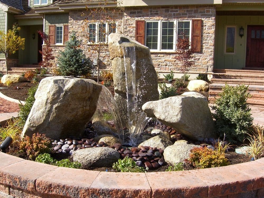Rock fountain surrounded by colorful plants and stone pathway in a landscaped yard.